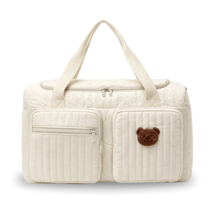 Cream diaper bag with quilted texture, zippered front pockets, and a brown teddy bear patch.