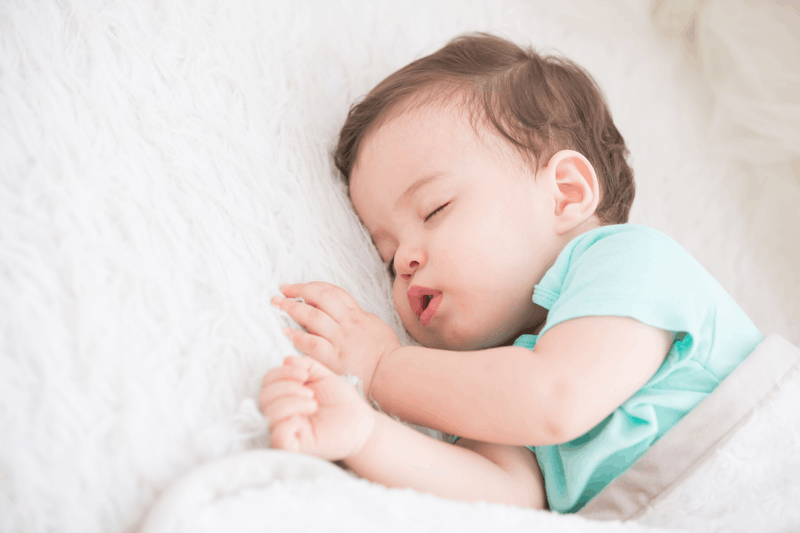 Baby sleeping peacefully on a white bed, wearing a light blue outfit.